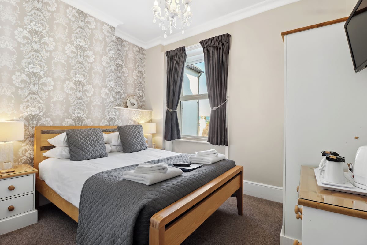 A neatly made bed with a quilted cover, a bedside table with a lamp, and a wardrobe with a television.