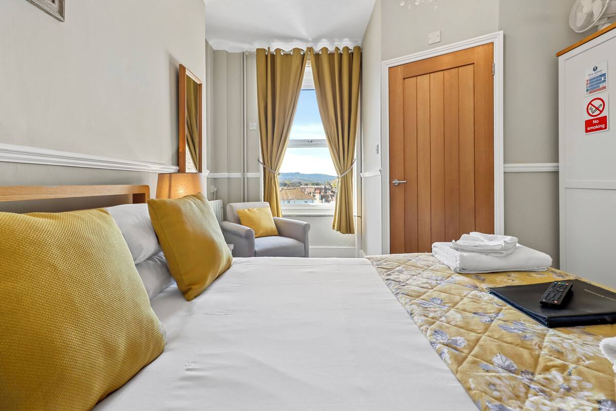 A hotel room with a bed, a chair, a desk with a laptop, a window with curtains, and a view of the city.