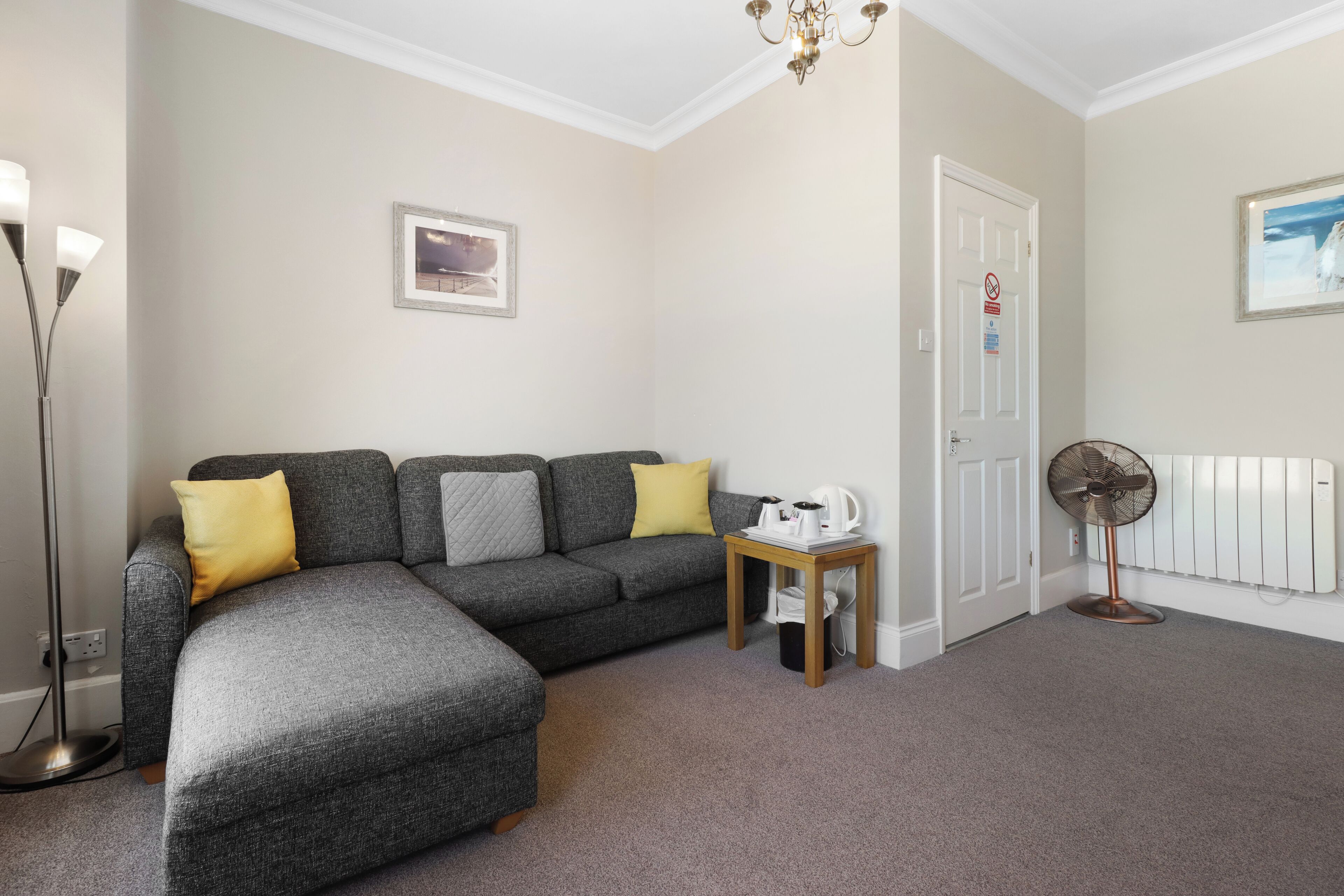 A living room with a grey sectional sofa, a small wooden table, a standing fan, and a radiator.