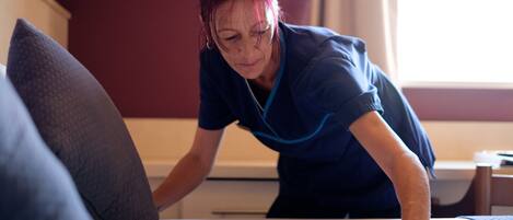 A person in a blue uniform cleaning a bed in a room with a red wall and a window.