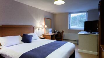 A hotel room with a large bed, a desk with a chair, a television, and a window with blinds.