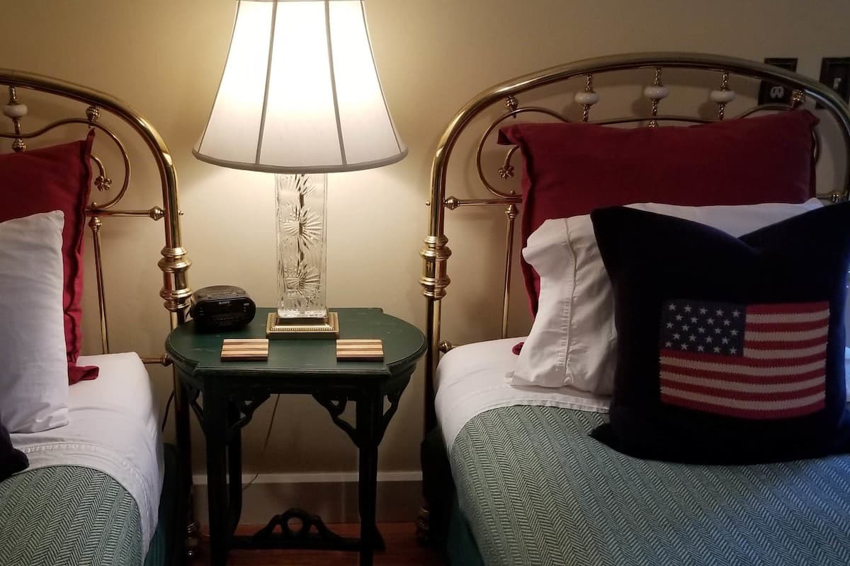 A hotel room with two beds, each with a pillow featuring an American flag design. A bedside table with a lamp and a small clock is between the beds.