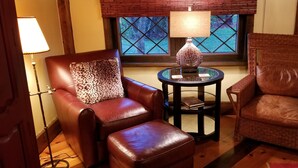 Una habitación acogedora con dos sillones de cuero, uno con un cojín de estampado de leopardo, una mesita de cristal con una lámpara, y vistas a través de una ventana con marco de madera.