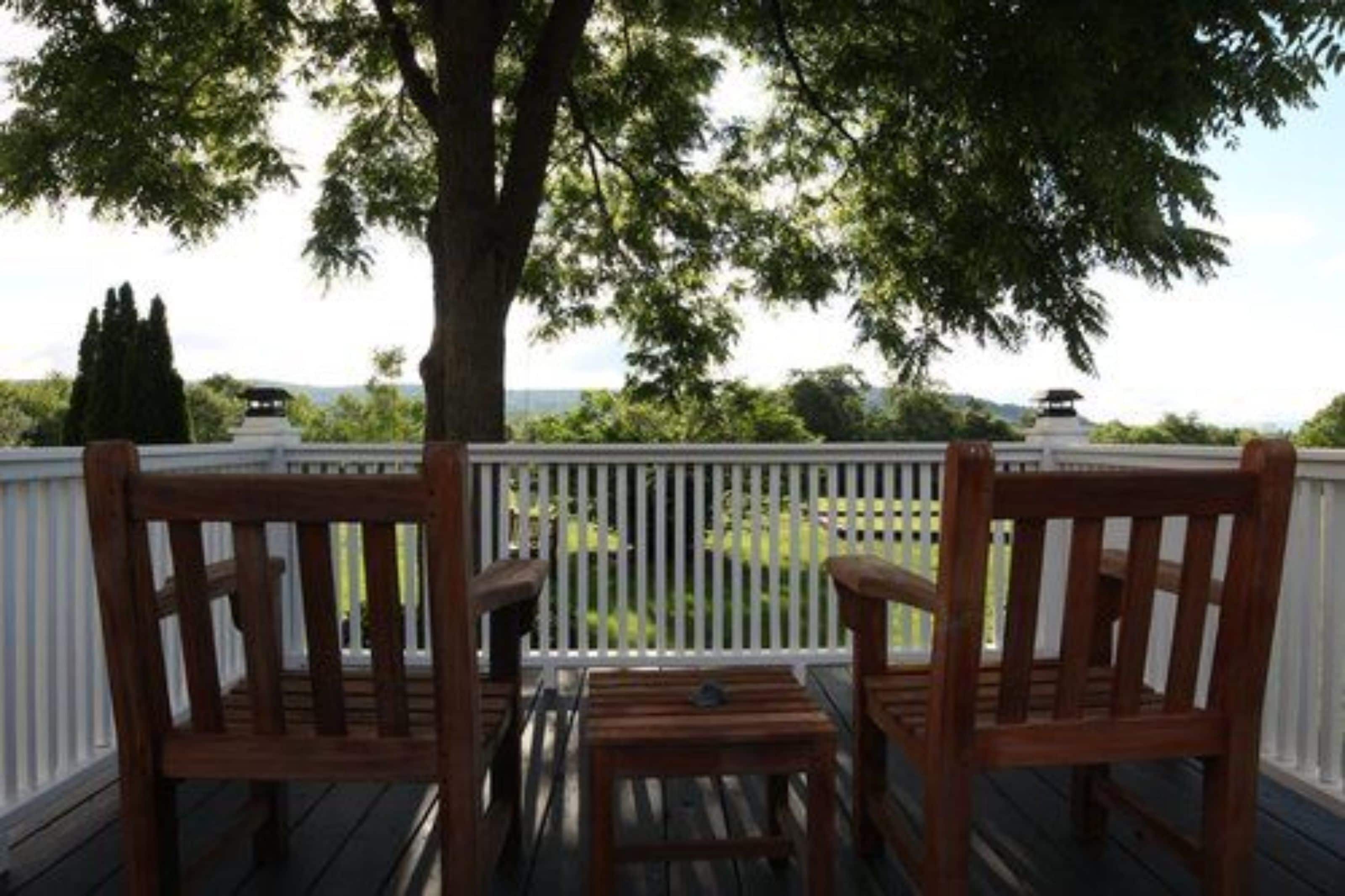 A wooden deck with two chairs and a small table, overlooking a green landscape with trees and a distant structure.