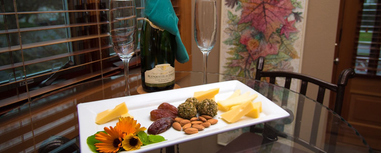 Una taula de vidre amb un plat blanc de formatge, almendres, dàtils i una flor. Es veuen també dos vasos de vi i una ampolla de vi.