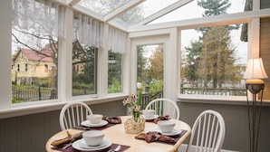 Ein Wintergarten mit Esstisch, gedeckt für vier Personen, umgeben von weißen Stühlen und einer Lampe auf der rechten Seite.