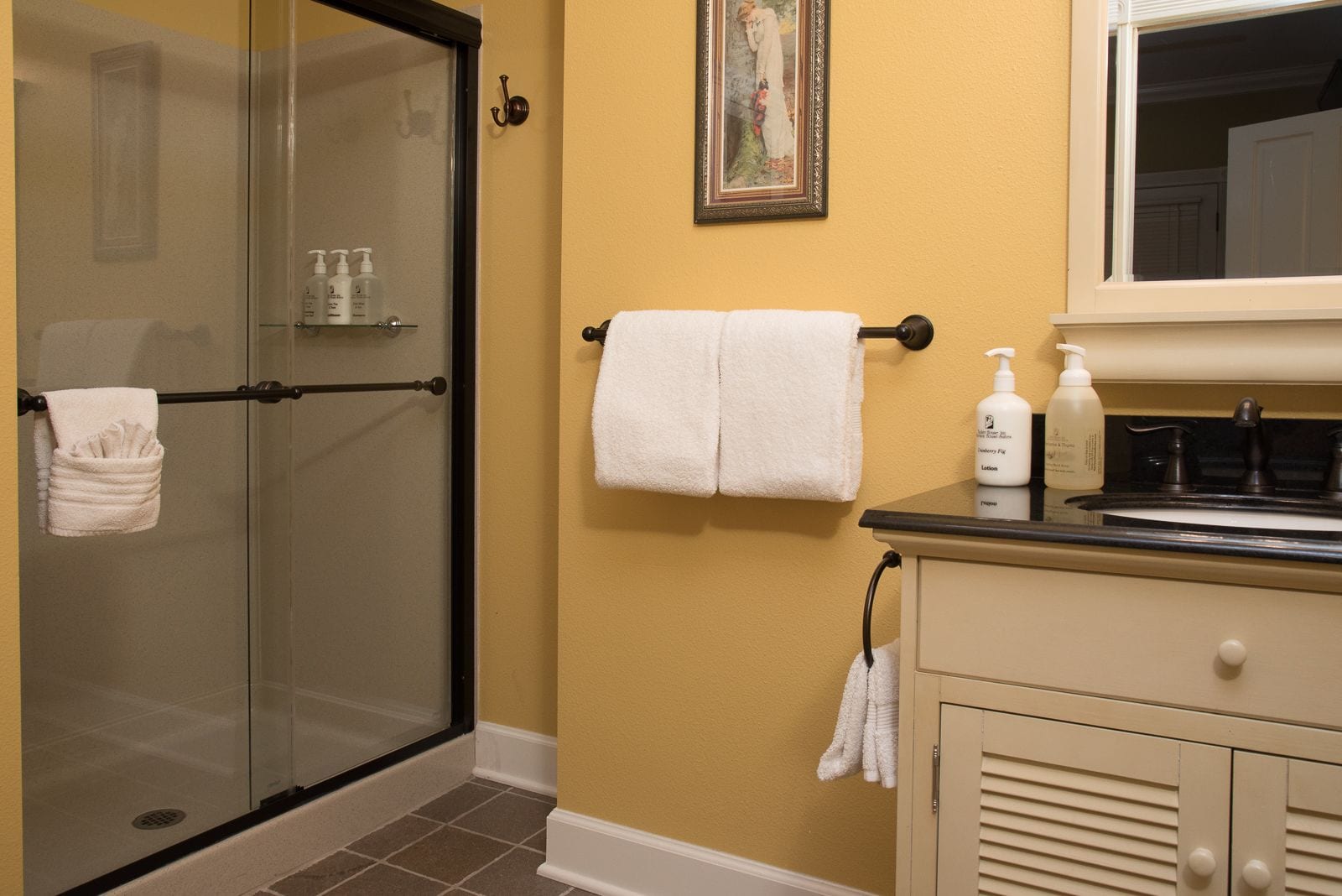 Un bagno con doccia in vetro, un portasciugamani con due asciugamani, un quadro appeso alla parete e articoli da toilette sul lavabo.