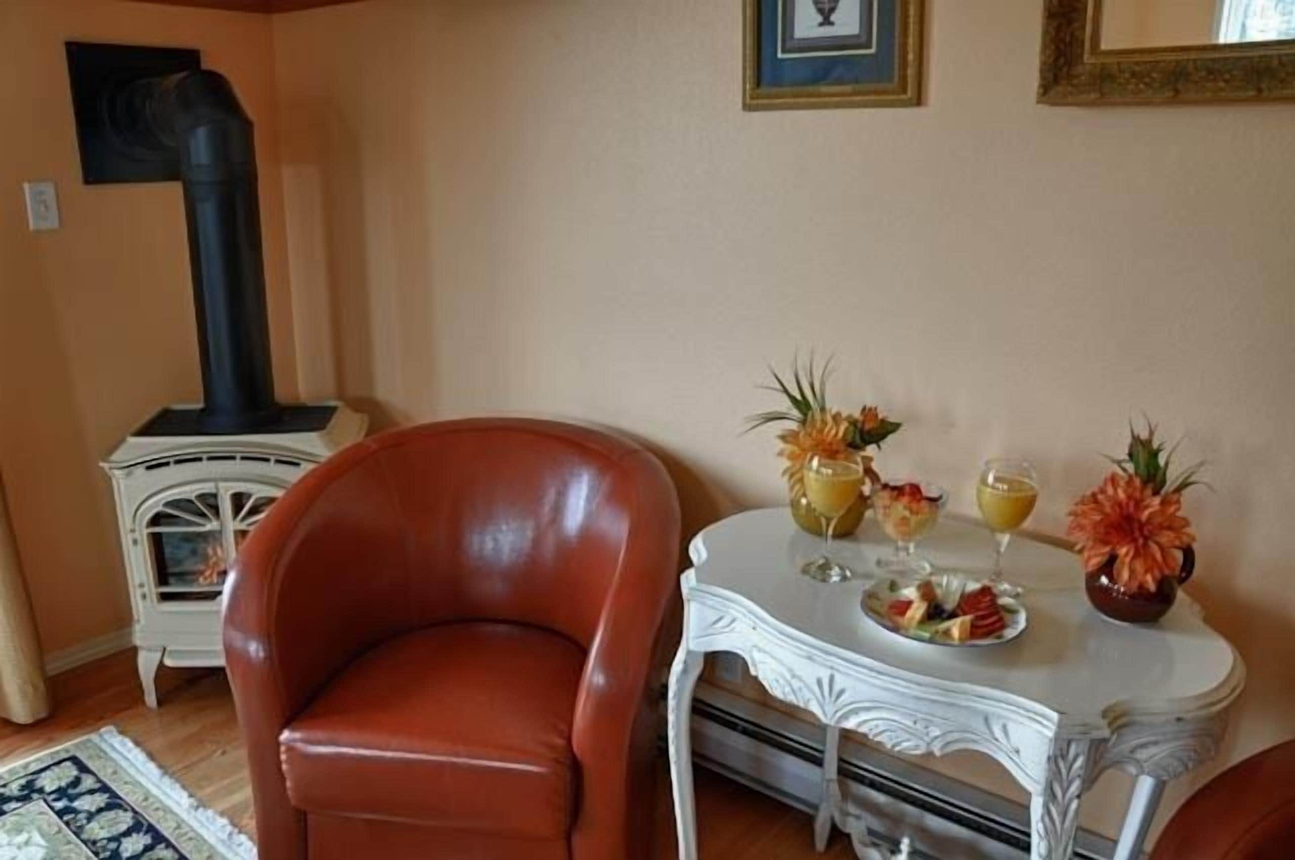 Un salon avec un fauteuil en cuir, une table blanche sur laquelle sont disposés des fruits et des boissons, une cheminée et un tableau encadré accroché au mur.