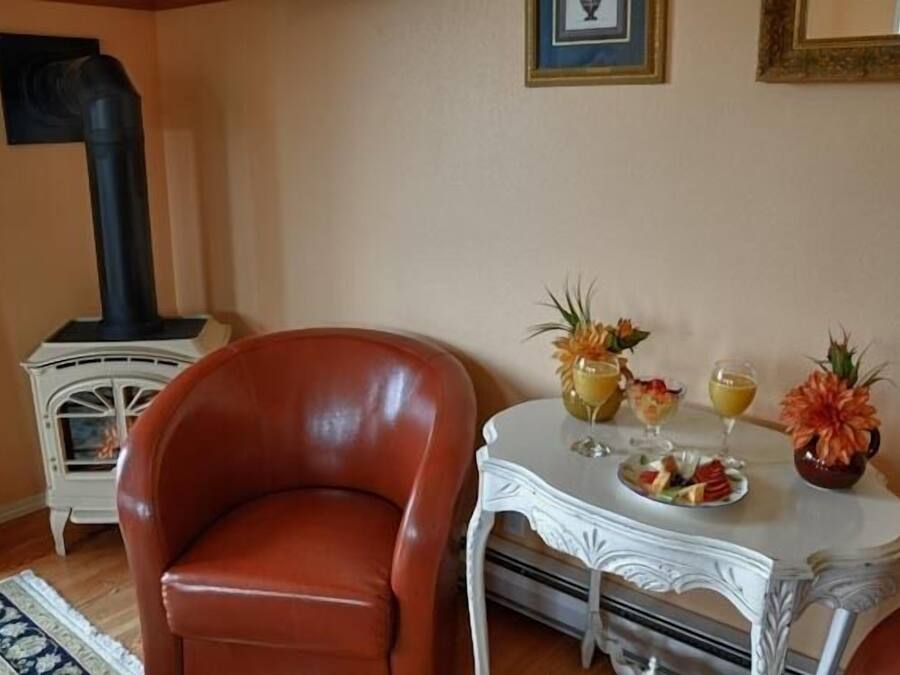 Un salon avec un fauteuil en cuir, une table blanche sur laquelle sont disposés des fruits et des boissons, une cheminée et un tableau encadré accroché au mur.