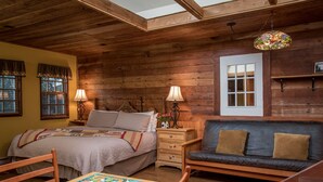 Ein gemütliches Zimmer mit Holzbalkendecke, einem Bett mit Steppdecke, einer Couch mit Kissen, einem Tisch mit einem Gesellschaftsspiel und einem Fenster mit Fensterläden.