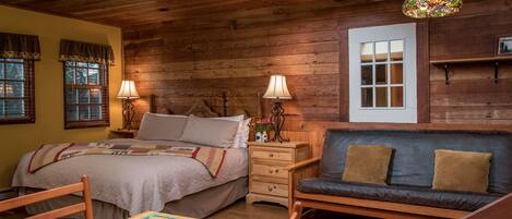 A cozy room with a wooden ceiling, a bed with a quilt, a sofa with cushions, a table with a board game, and a window with shutters.