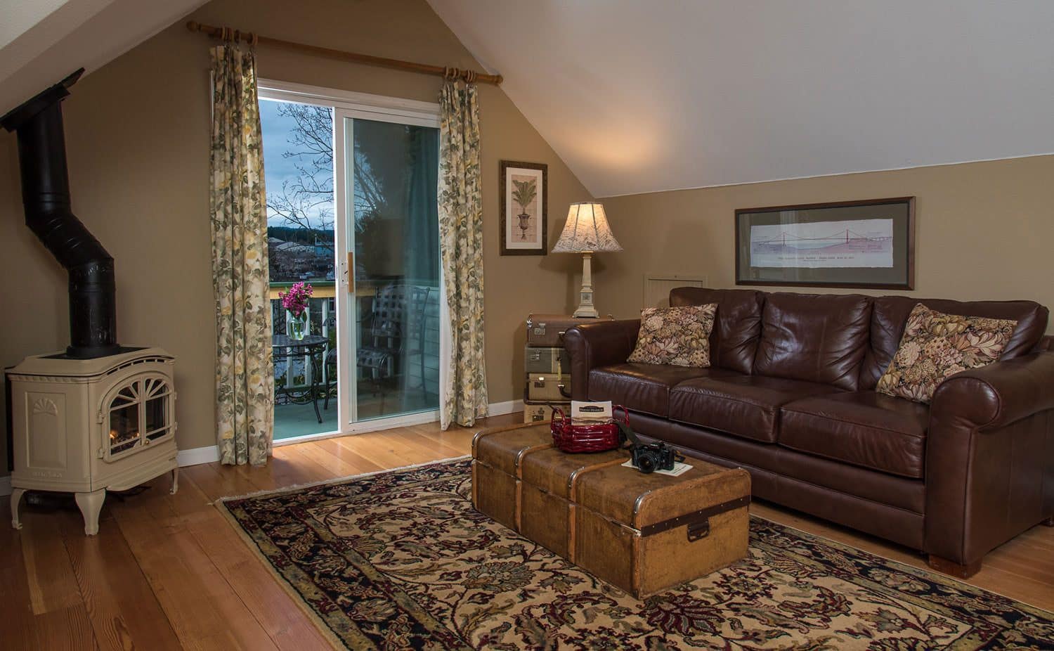 A cozy living room with a brown leather sofa, a vintage suitcase, a wood-burning stove, and a large window with curtains.