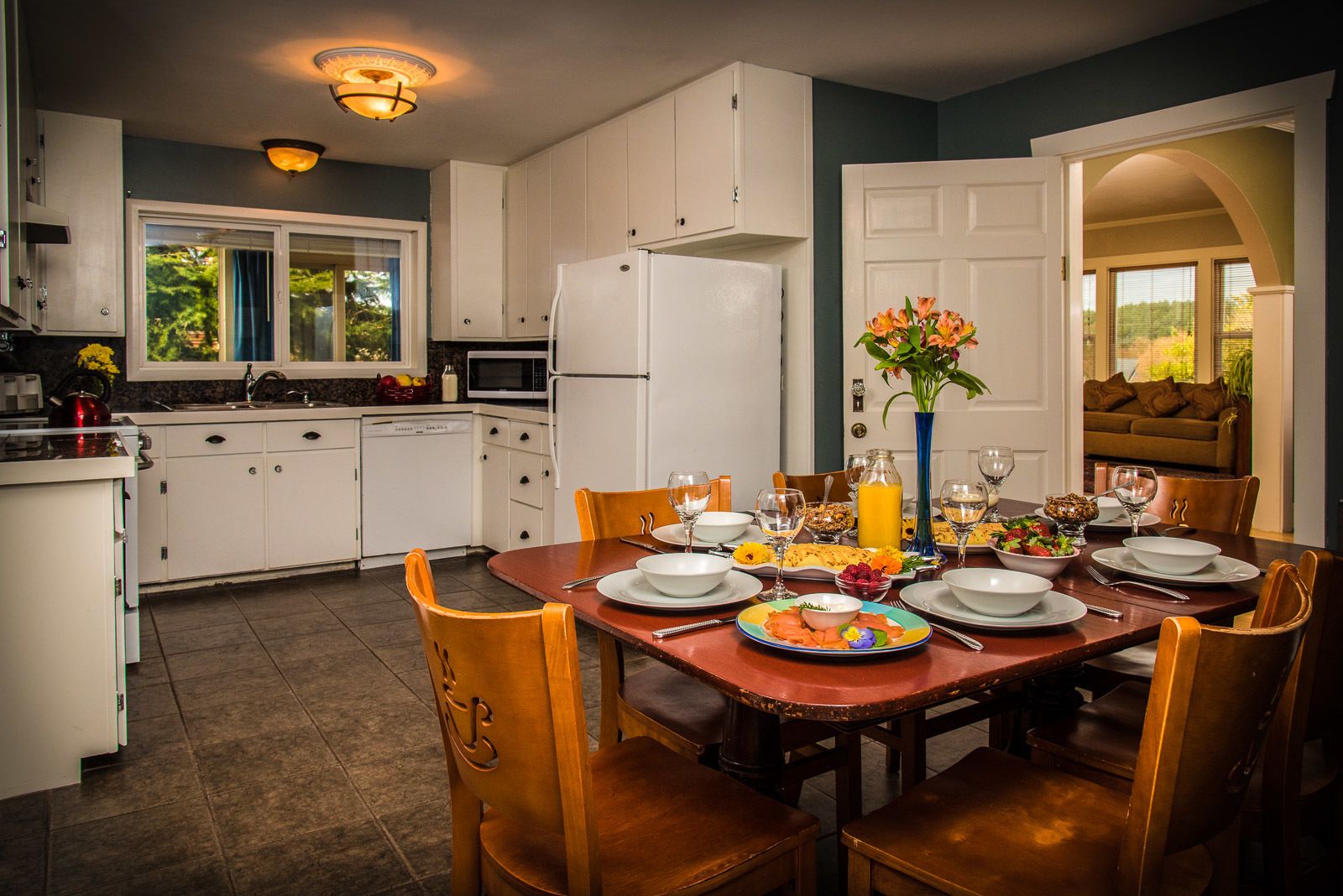 A modern kitchen with white cabinets, a dining table set for four, and a view into a living area with a sofa.