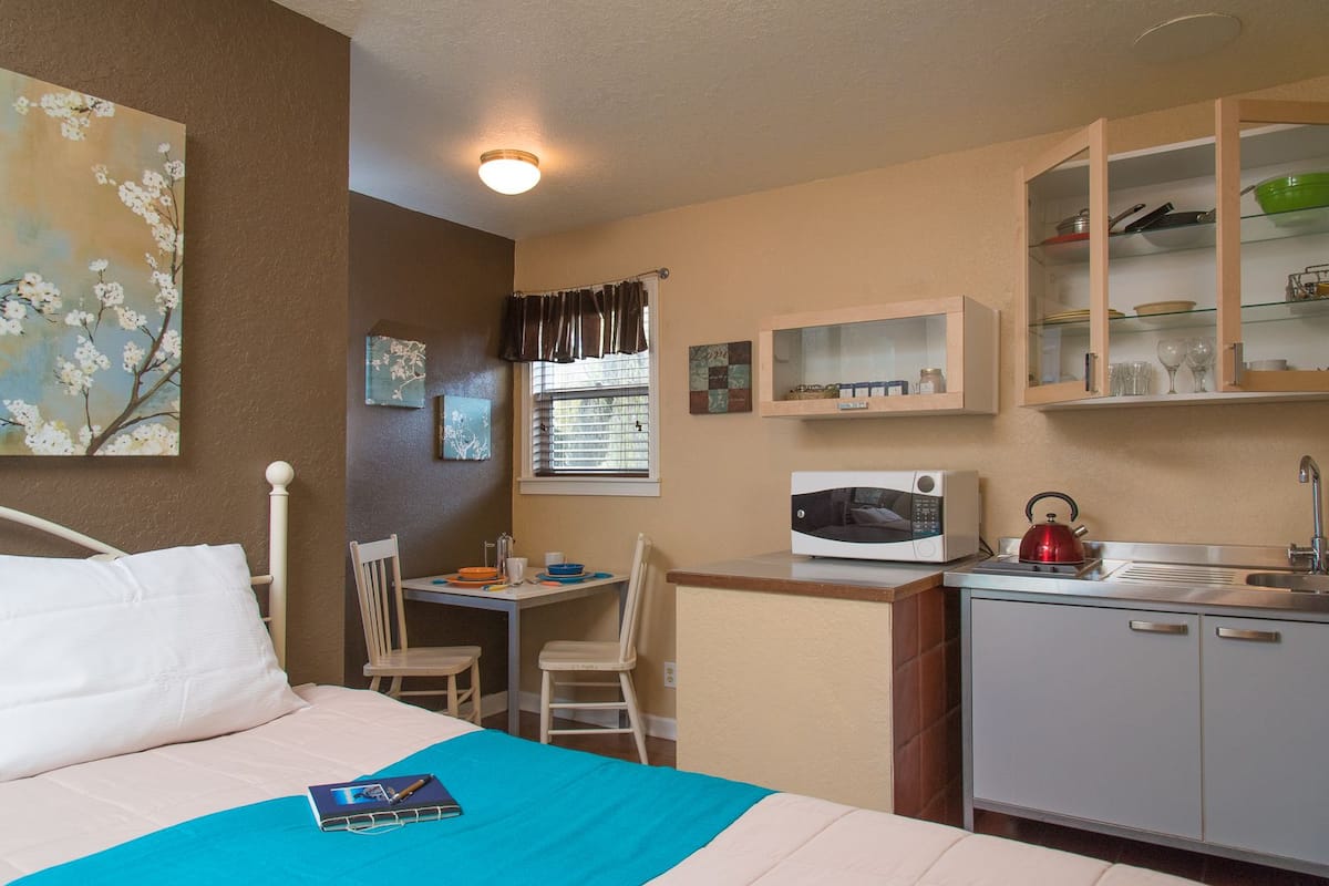 Une chambre d’hôtel compacte comprenant un lit, un petit bureau avec une chaise, un four à micro-ondes, une bouilloire et une kitchenette équipée d’armoires et d’étagères.
