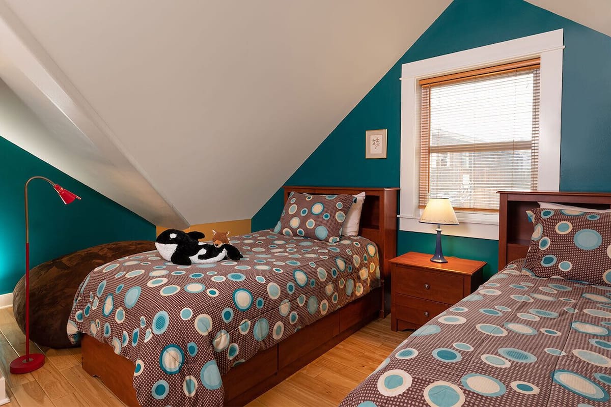 Une chambre avec deux lits, une fenêtre avec des stores, une table de chevet et une lampe.