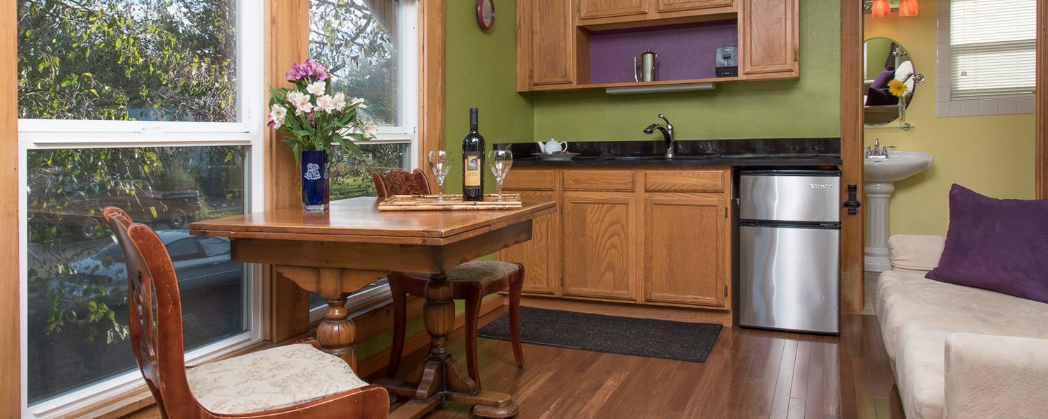 A kitchen with wooden cabinets, a stainless steel refrigerator, a dining table with chairs, and a window with a view of trees.