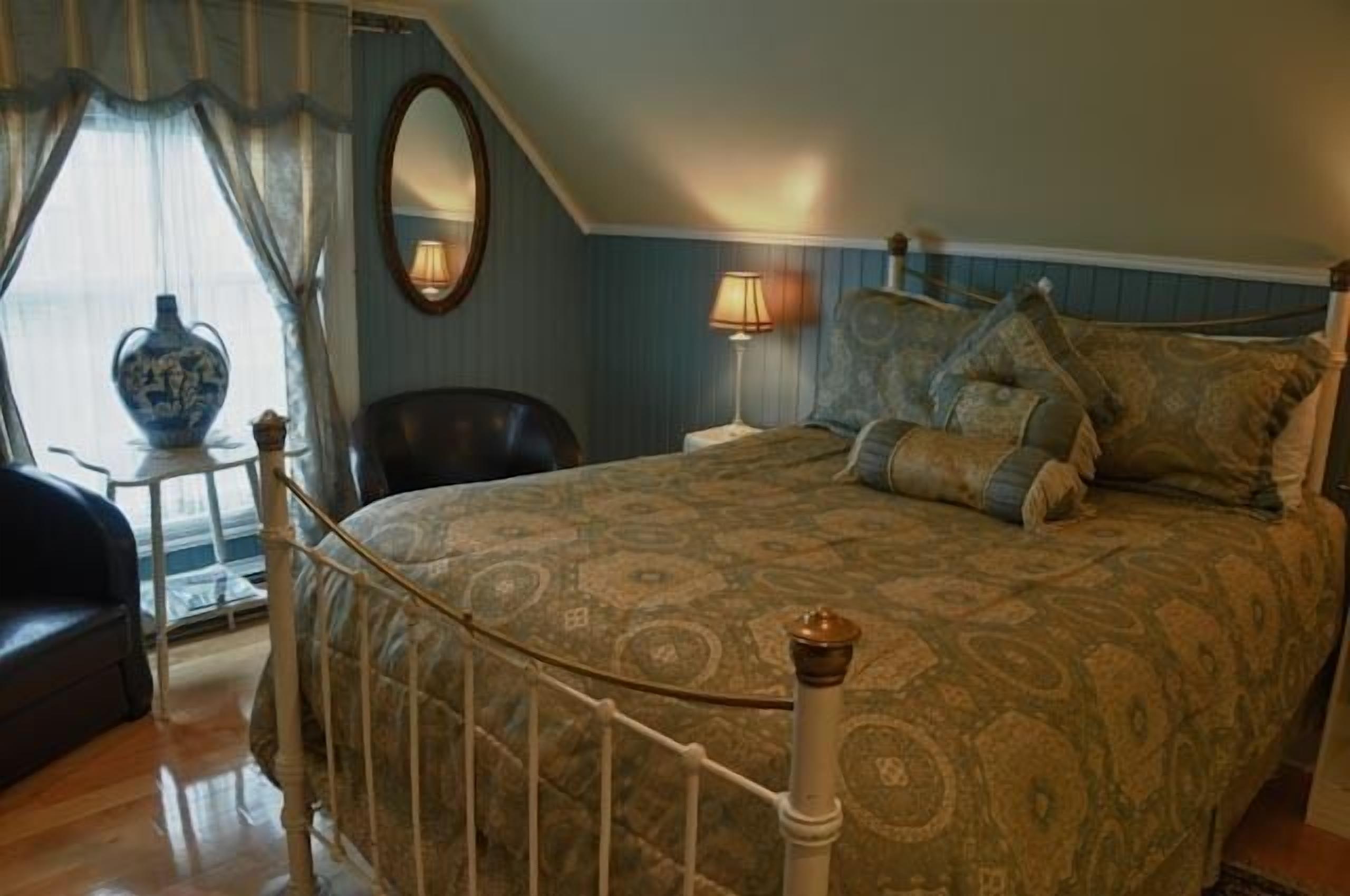 A bedroom with a patterned bedspread, a round mirror, a bedside lamp, and a chair.