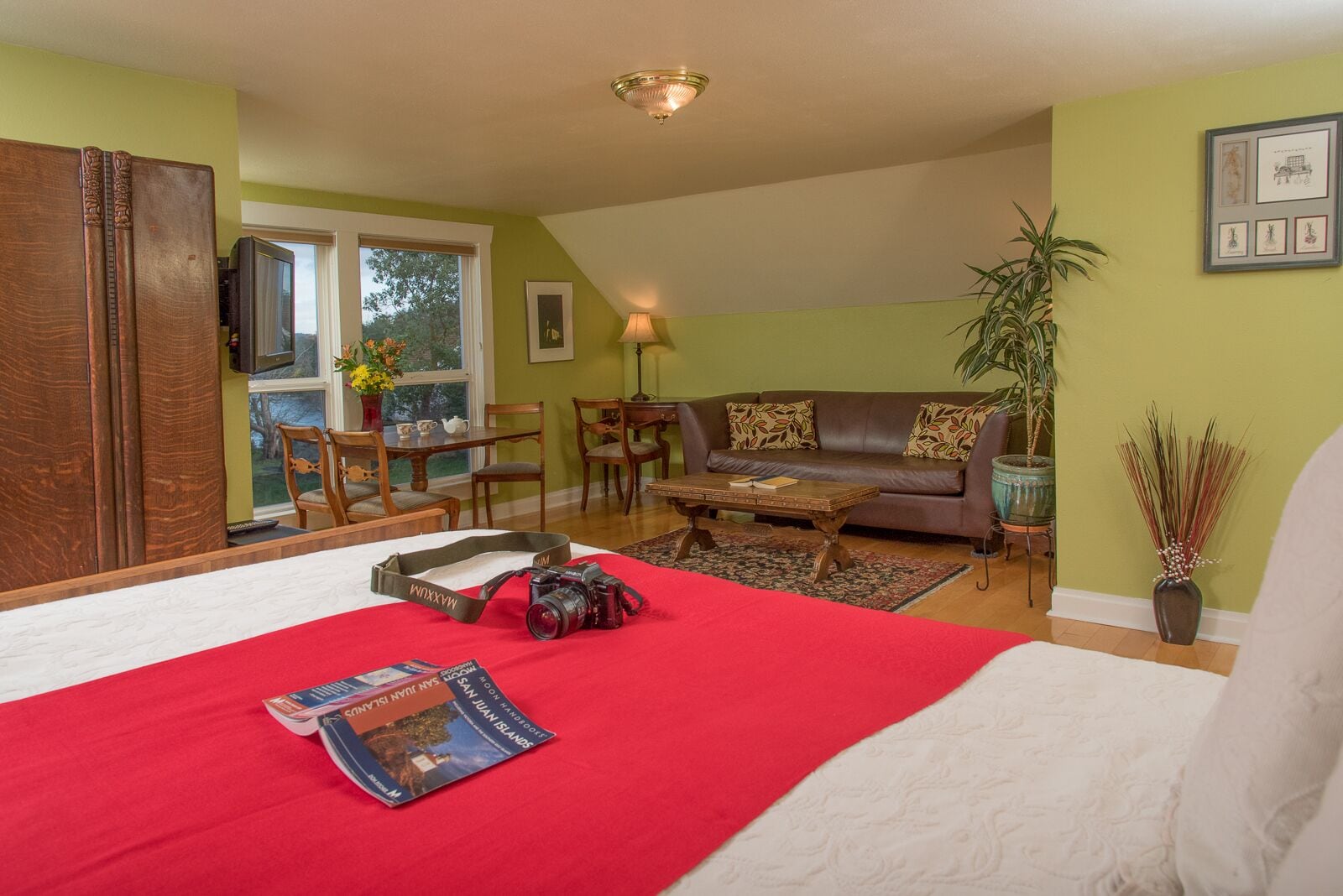 A cozy living room with a large bed, a brown leather sofa, a coffee table, and a dining area with wooden furniture.