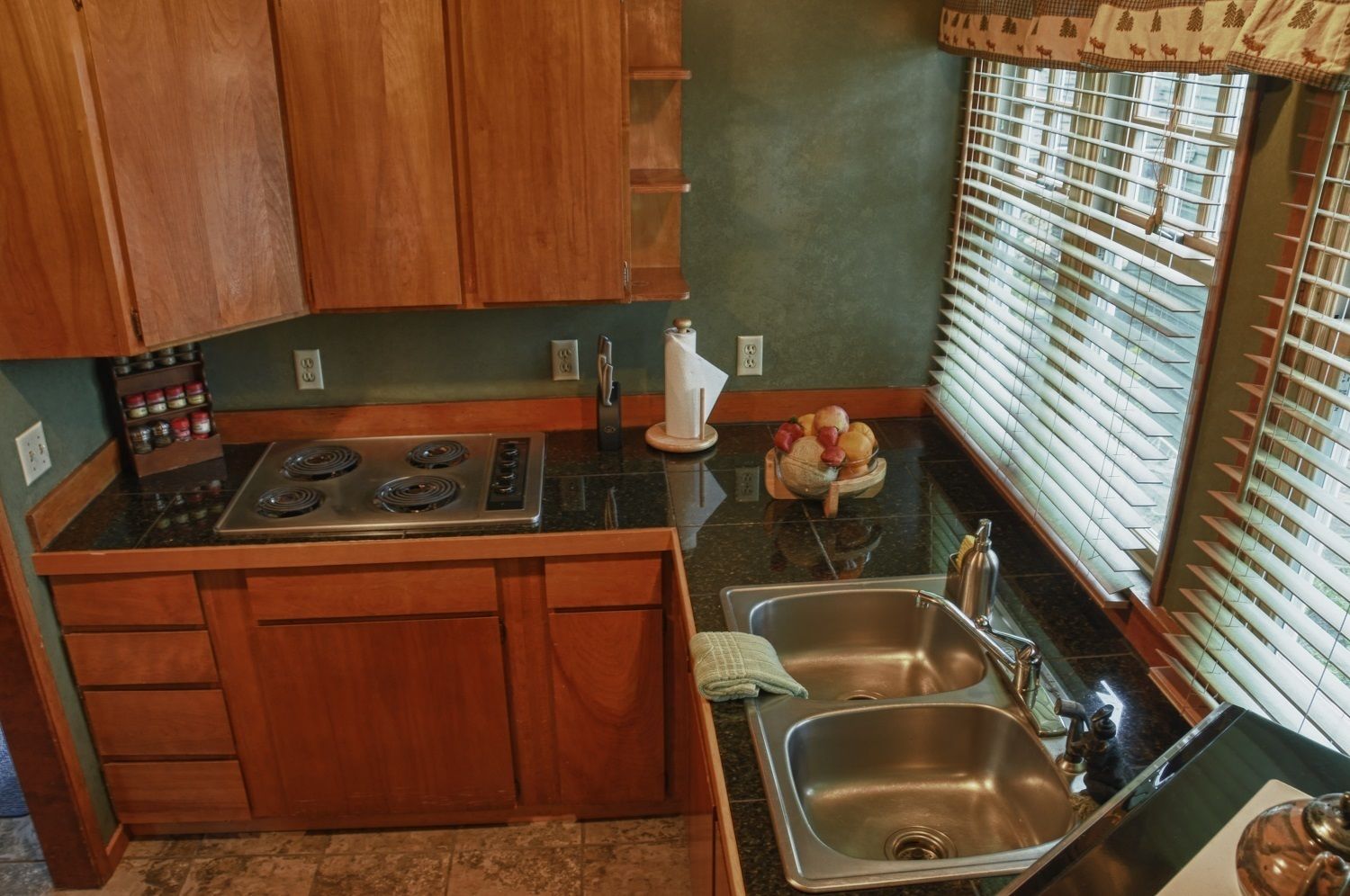 A compact kitchen with wooden cabinets, a stove, a sink, and a window with blinds.
