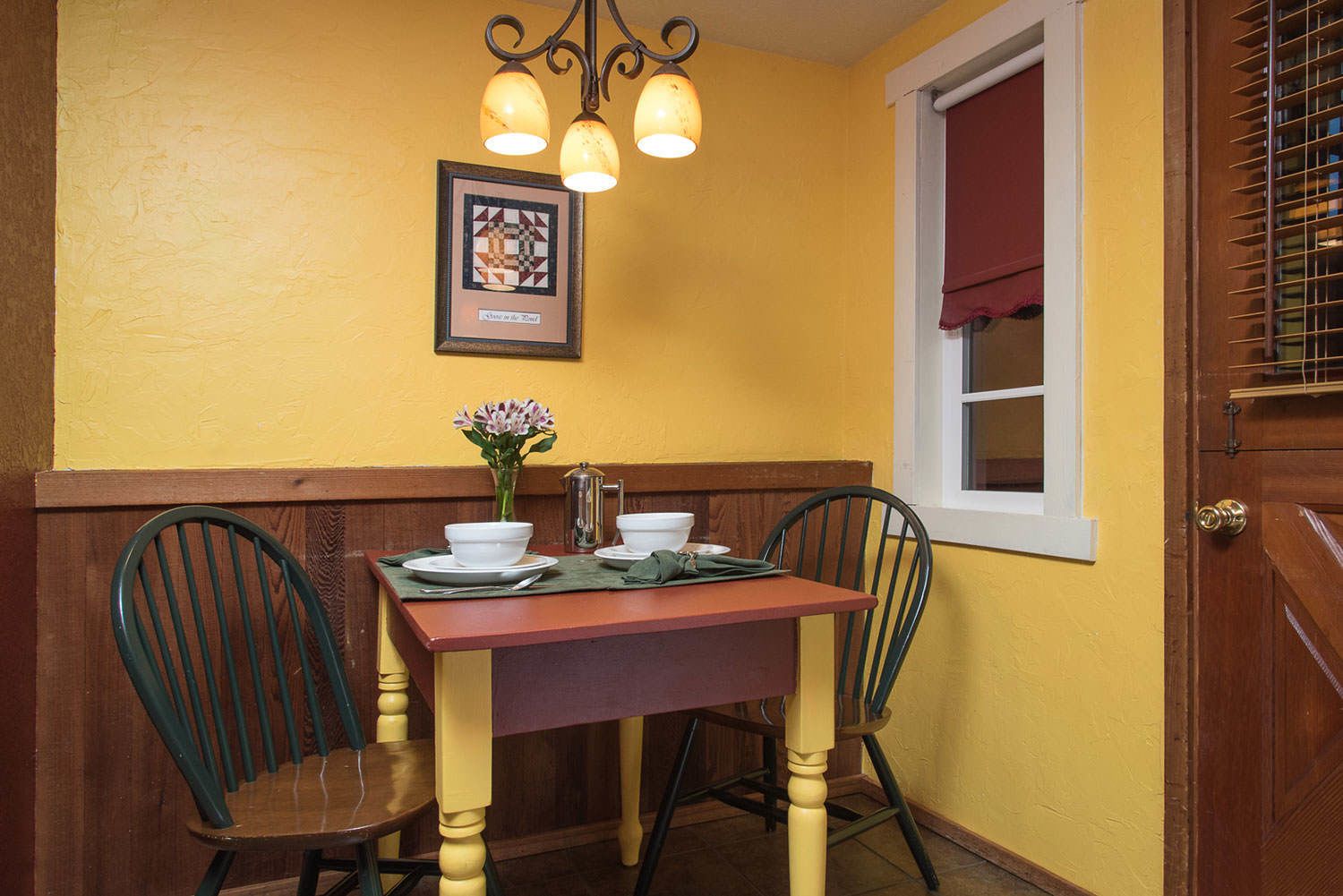 Un comedor con una mesa de madera, dos sillas, un florero con flores y un cuadro en la pared.