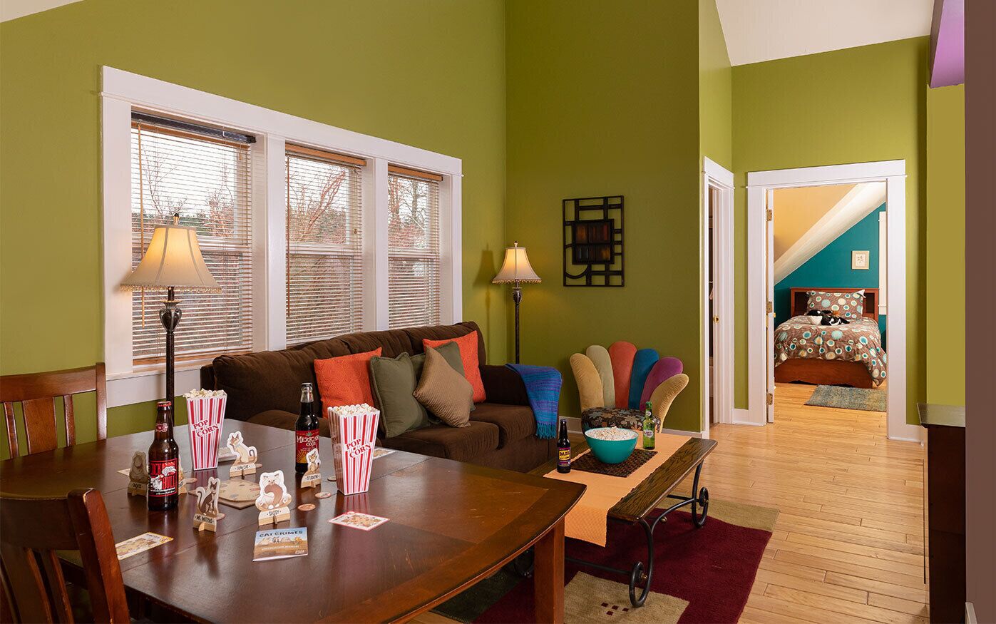 A cozy living room with a wooden dining table, popcorn, and a bottle of beer. A brown couch with colorful cushions is in the background.