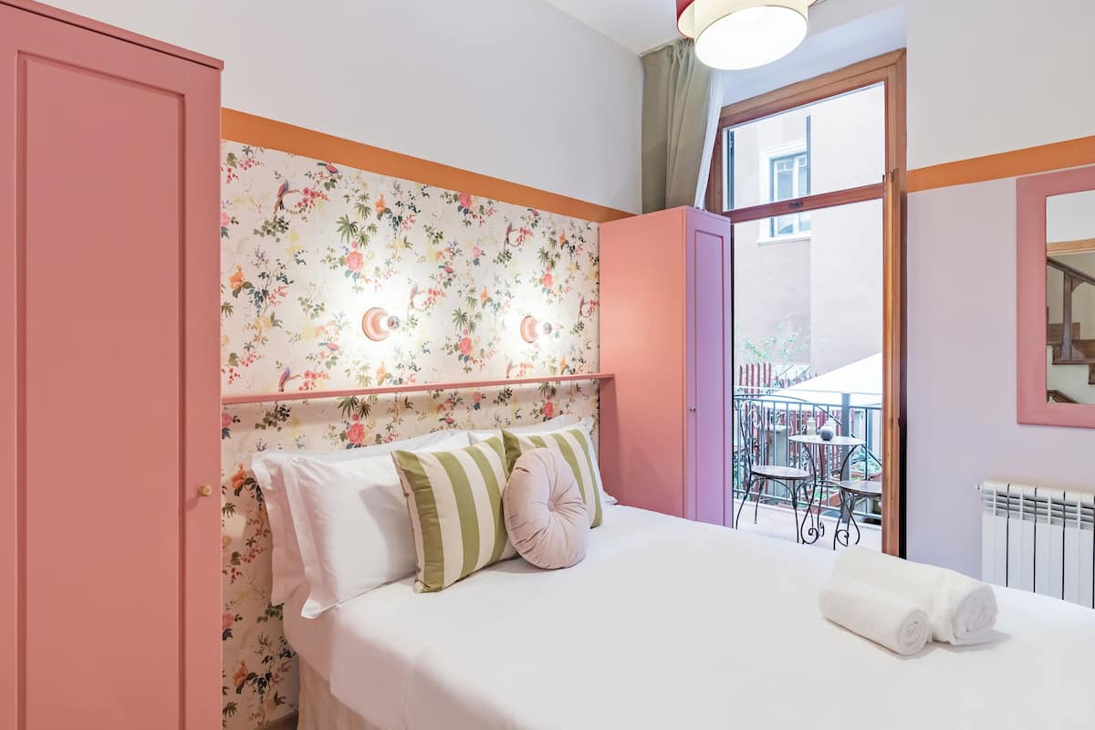 A bedroom with a floral wallpaper, a bed with white bedding and striped pillows, a pink door, and a balcony with a table and chairs.