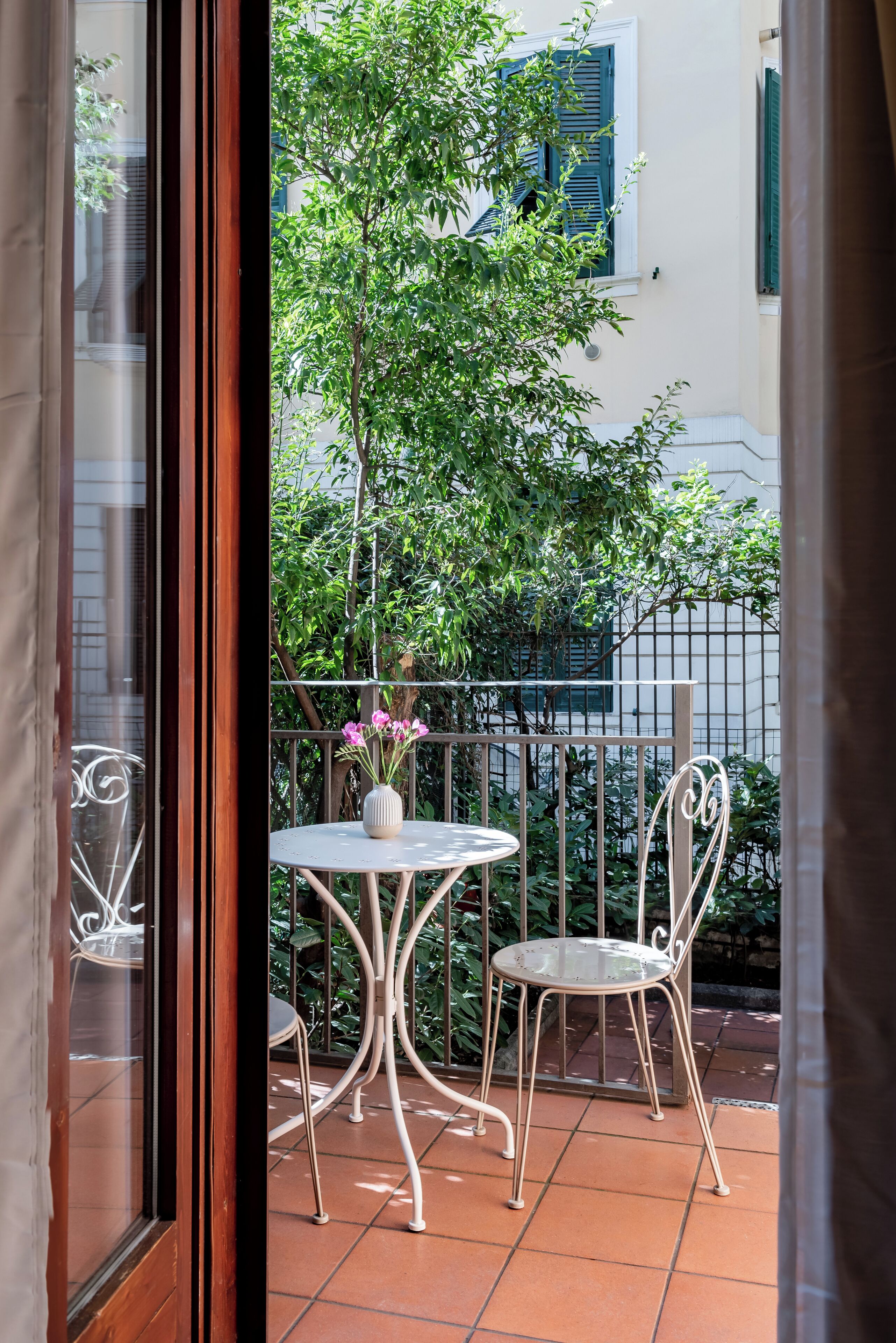 A balcony with a round table and two chairs, a potted plant, and a view of a tree and building.