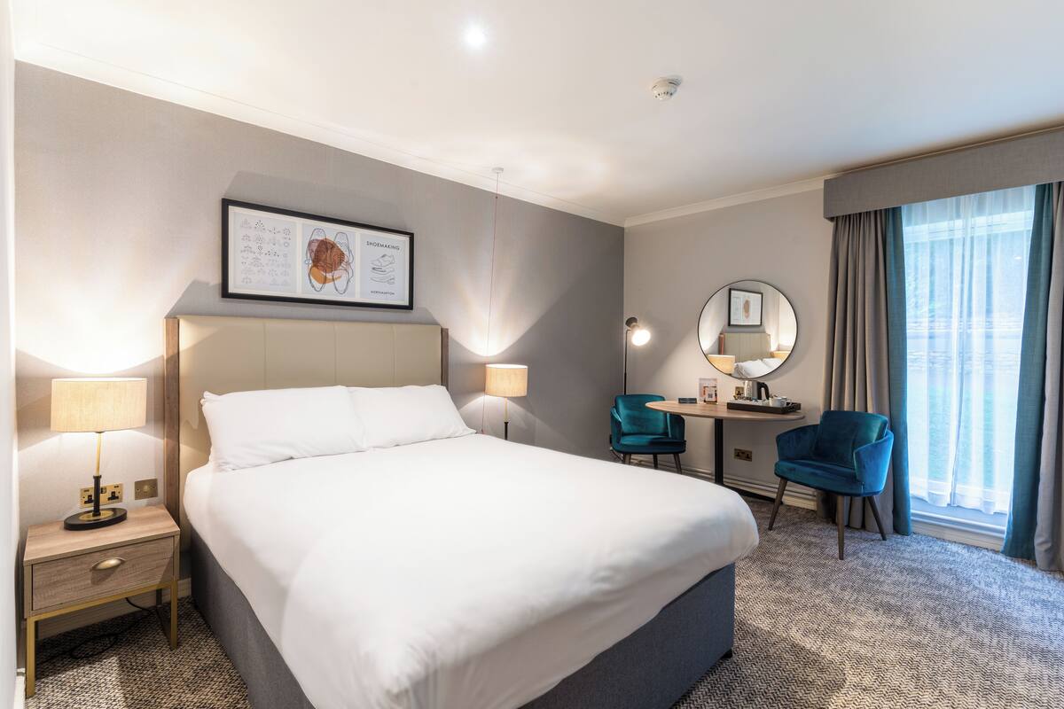 A hotel room with a large bed, bedside lamps, a desk with a chair, a mirror, and a window with curtains.