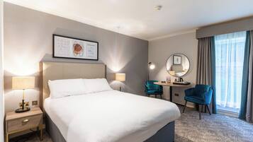 Habitación de hotel con una cama grande, lámparas de noche, un escritorio con silla, un espejo y una ventana con cortinas.