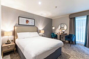 Habitación de hotel con una cama grande, lámparas de noche, un escritorio con silla, un espejo y una ventana con cortinas.