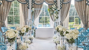 Une allée bordée de chaises et de vases remplis de fleurs, des rideaux élégamment drapés et une vue sur la verdure à l’extérieur.