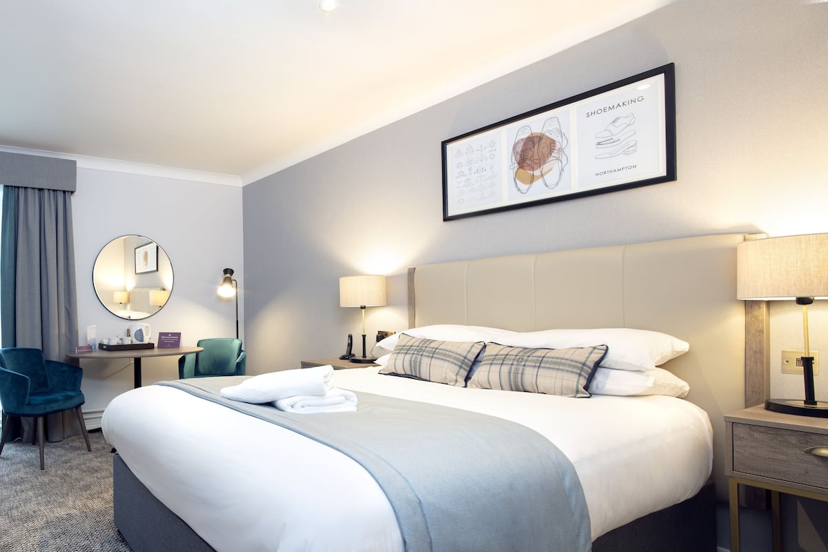 A hotel room with a large bed, a desk, a chair, a round mirror, and a framed picture on the wall.