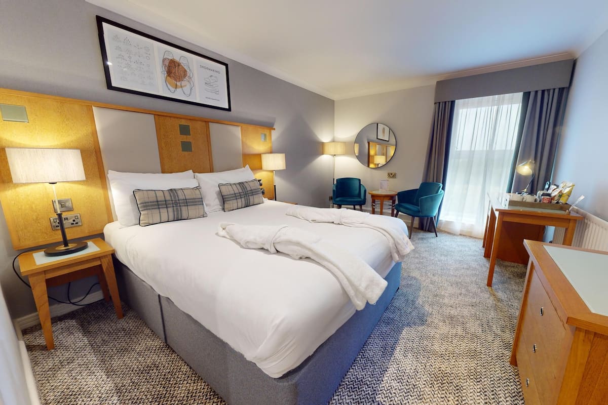 A hotel room with a large bed, two bedside tables with lamps, a desk with a chair, a blue armchair, a round mirror, and a window with curtains.