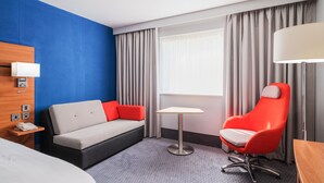 Une chambre d’hôtel avec un lit, un canapé, une petite table, une chaise rouge, une lampe et une fenêtre avec des rideaux.