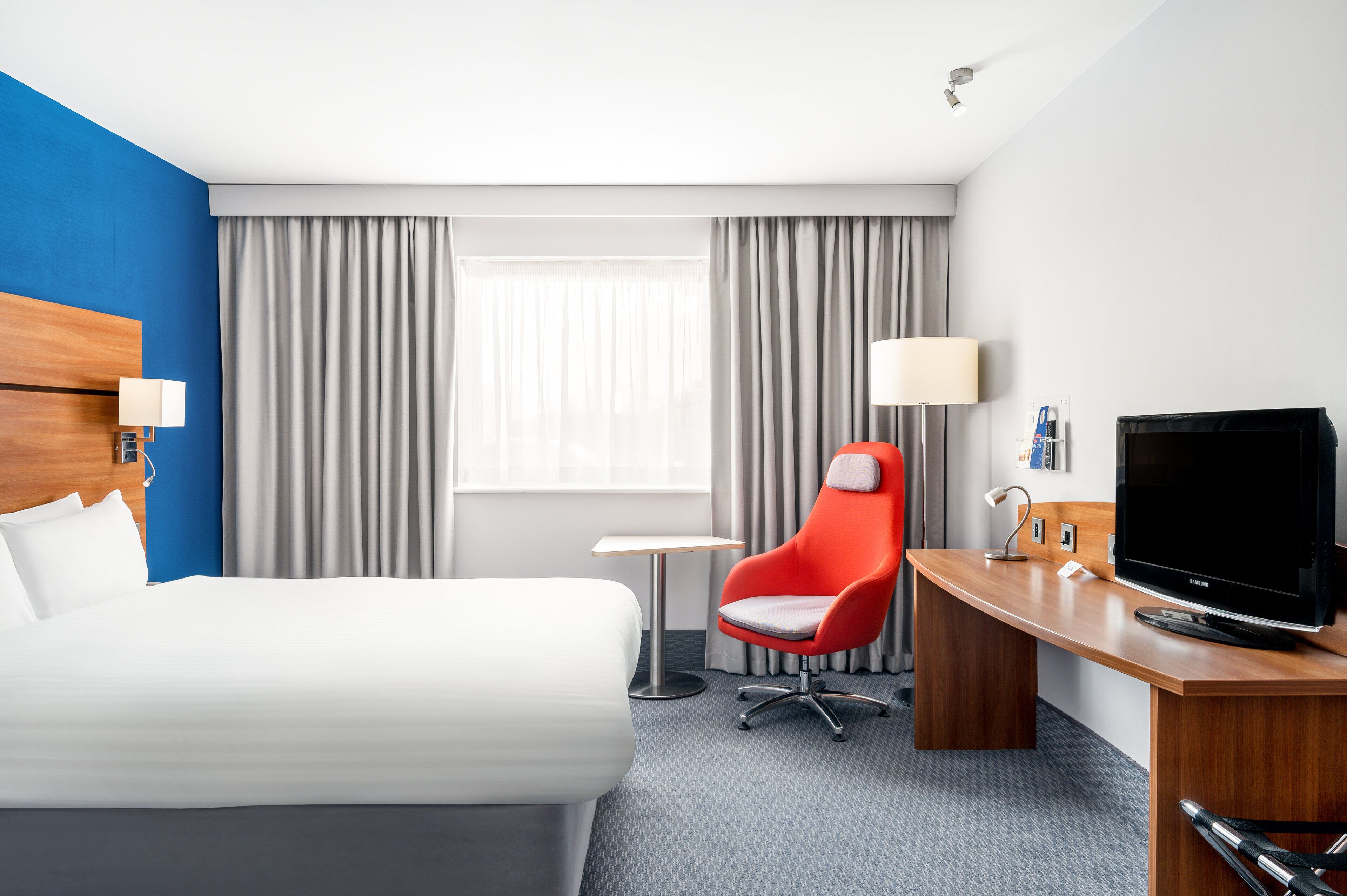 A hotel room with a large bed, a desk with a TV, a red chair, and a small table.