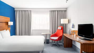 A hotel room with a large bed, a desk with a TV, a red chair, and a small table.