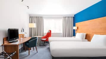 A hotel room with a bed, a desk with a TV, a chair, and a window with curtains.