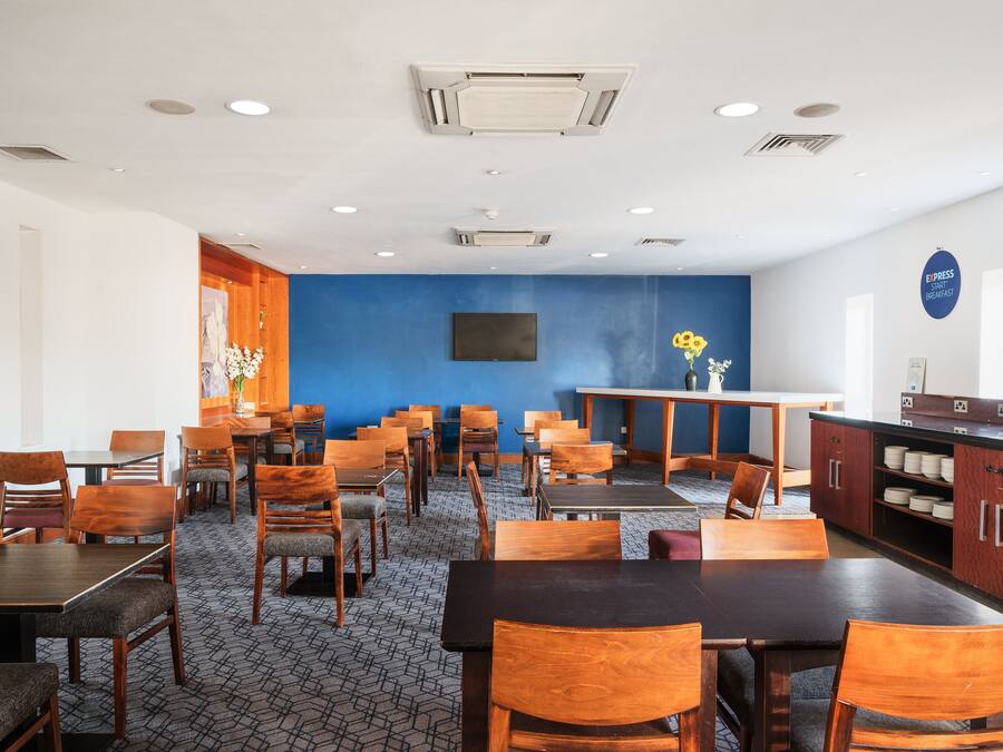 A hotel dining area with multiple tables and chairs, a blue accent wall, and a kitchen area with a sign reading "EXPRESS START YOUR BREAKFAST".