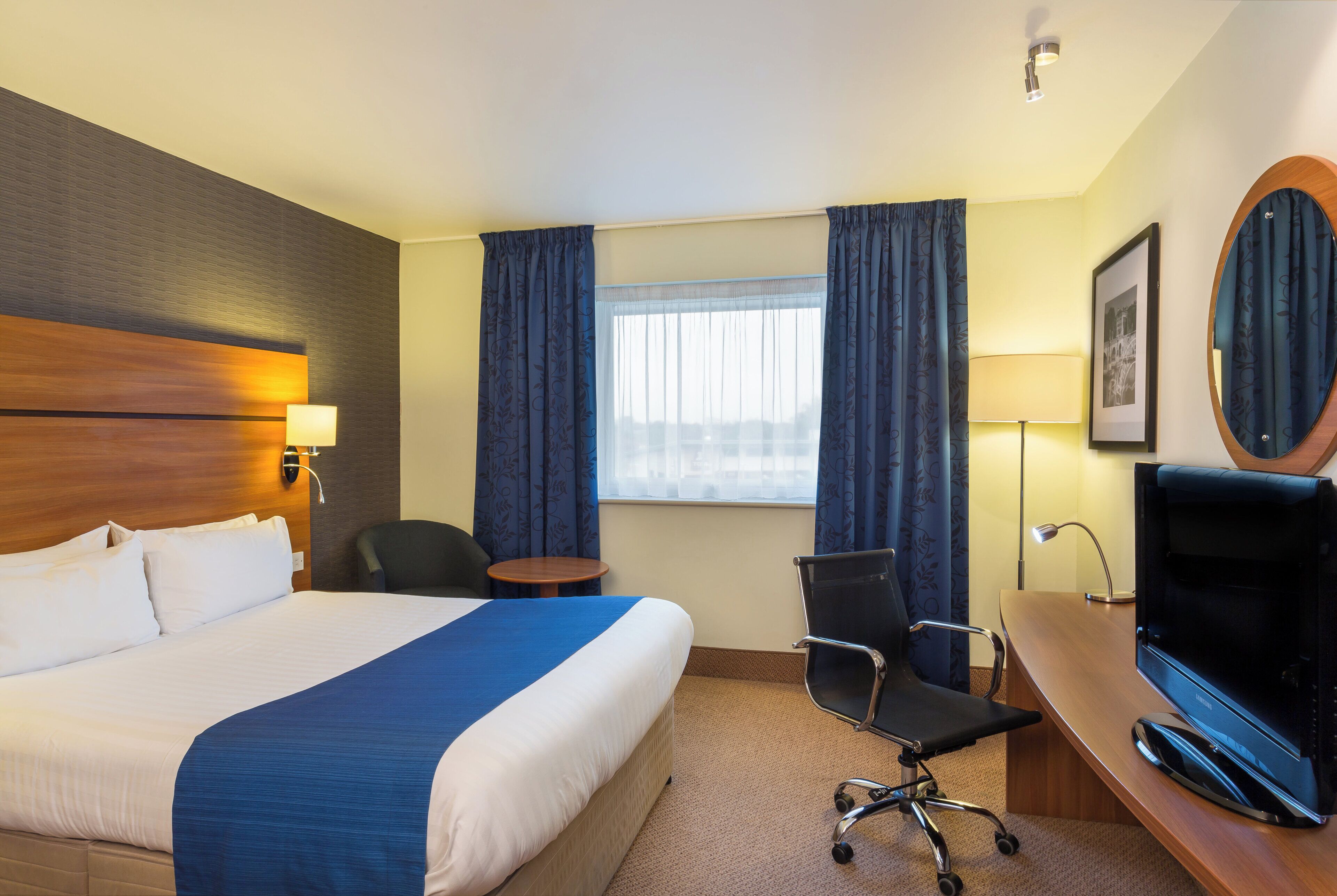 A hotel room with a large bed, a desk with a television, a chair, a small table, and a window with curtains.