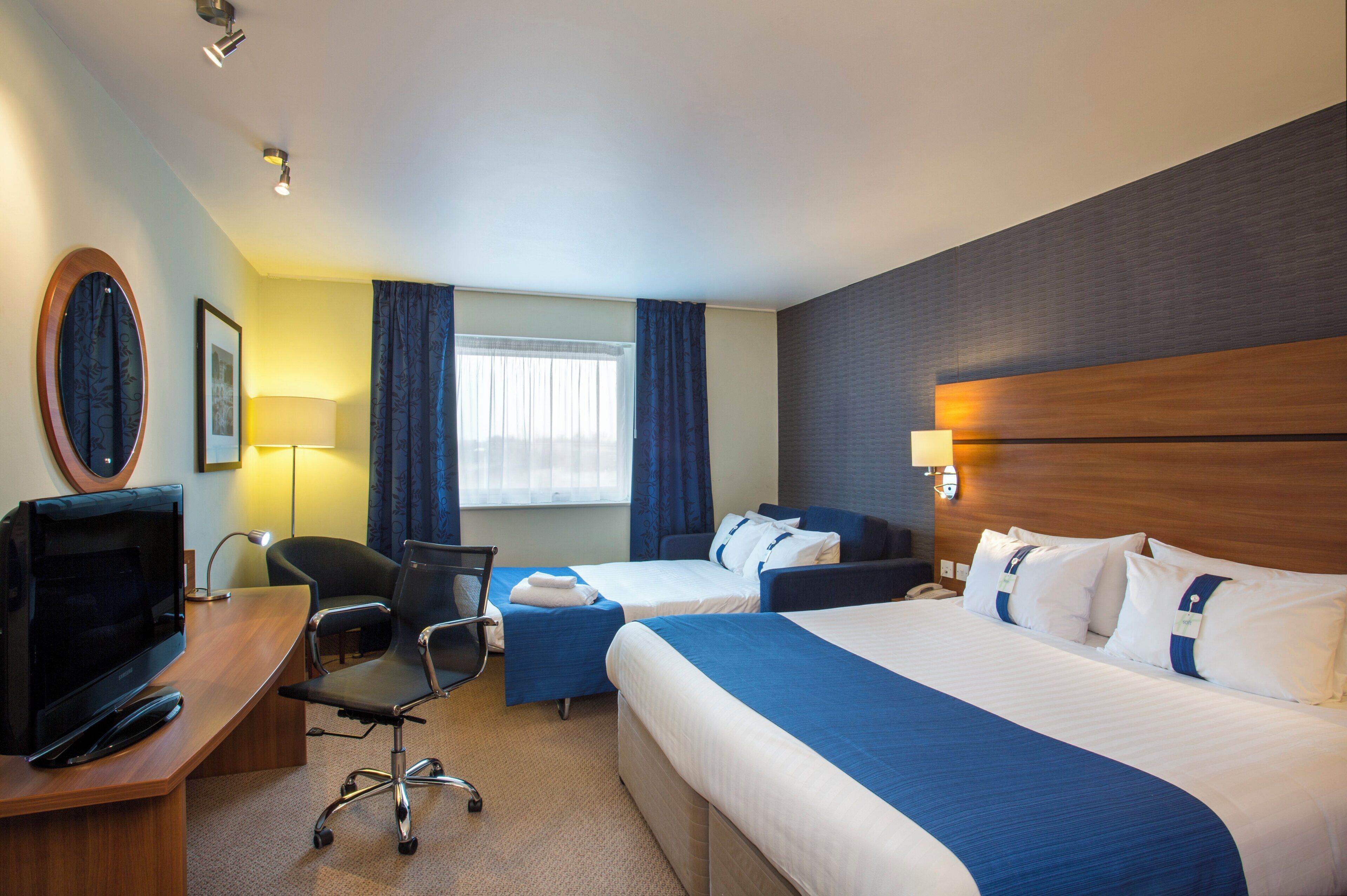 A hotel room with a desk, two beds, a TV, and a window with curtains.