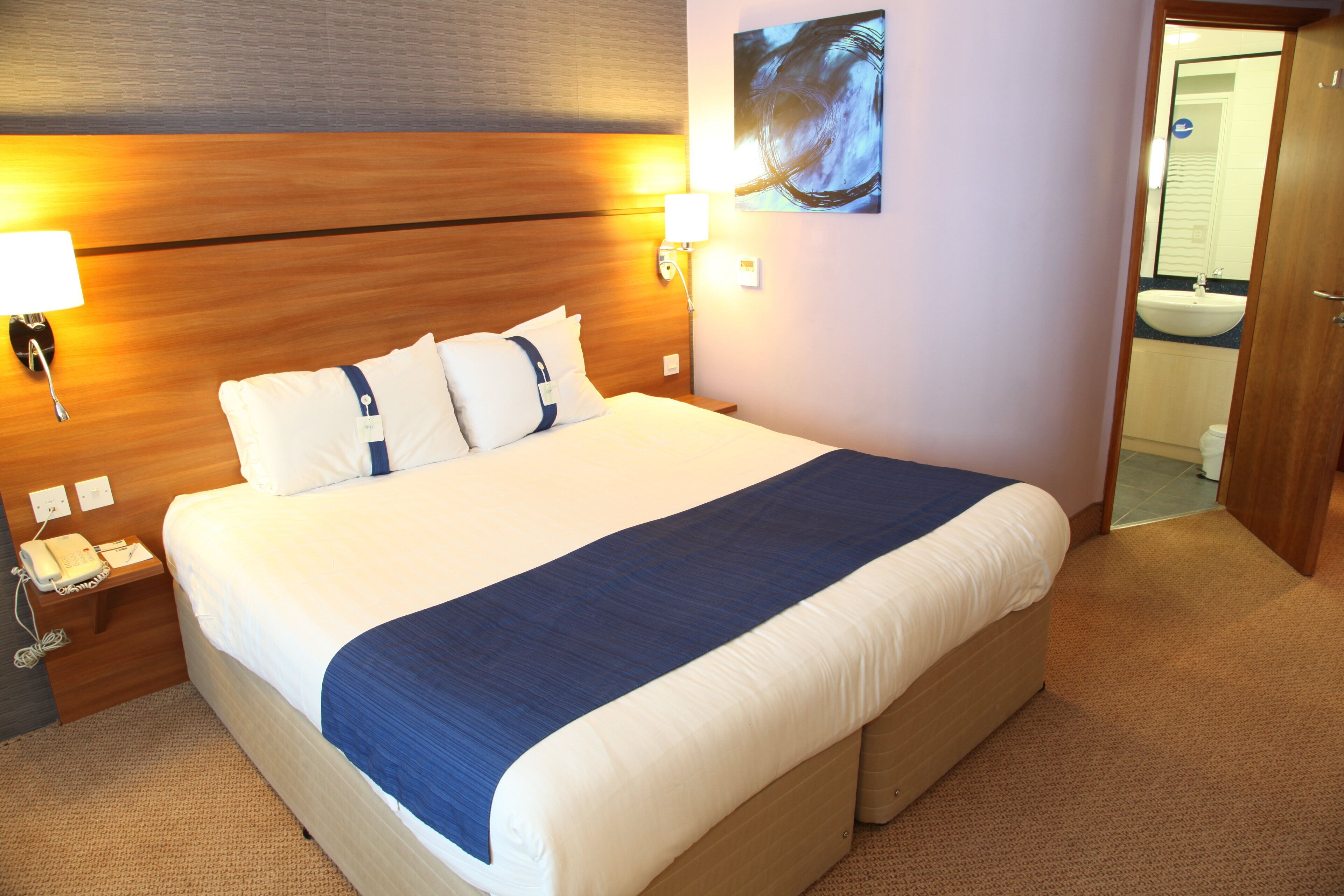 A hotel room with a bed, bedside lamps, a telephone, and a painting on the wall.