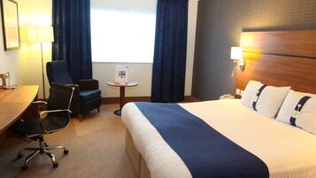 Habitación de hotel con una cama grande, un escritorio con silla, una mesita y un ventanal con cortinas.