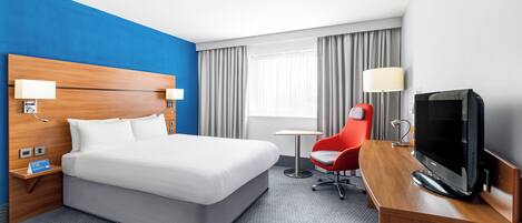 A hotel room with a bed, a desk with a TV, a chair, and a window with curtains.