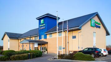 A Holiday Inn Express hotel with a blue and beige exterior, a parking lot with several cars, and a flagpole.