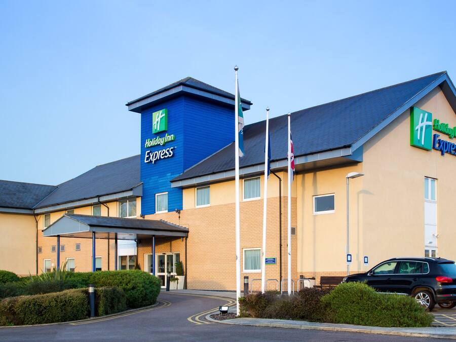 A Holiday Inn Express hotel with a blue and beige exterior, a parking lot with several cars, and a flagpole.