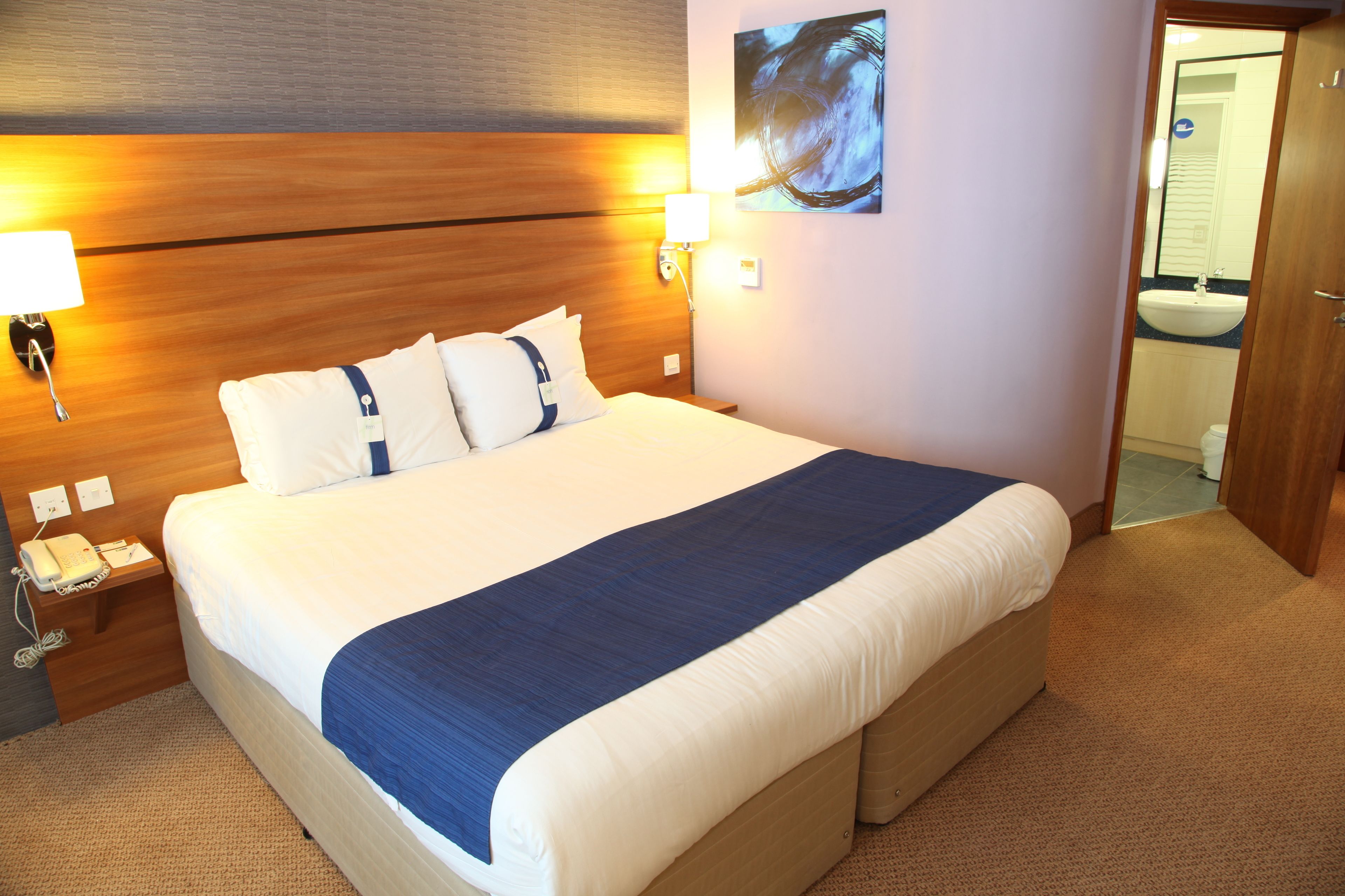 Una habitación de hotel con una cama grande, una mesita de noche con una lámpara, un teléfono y un cuadro en la pared.