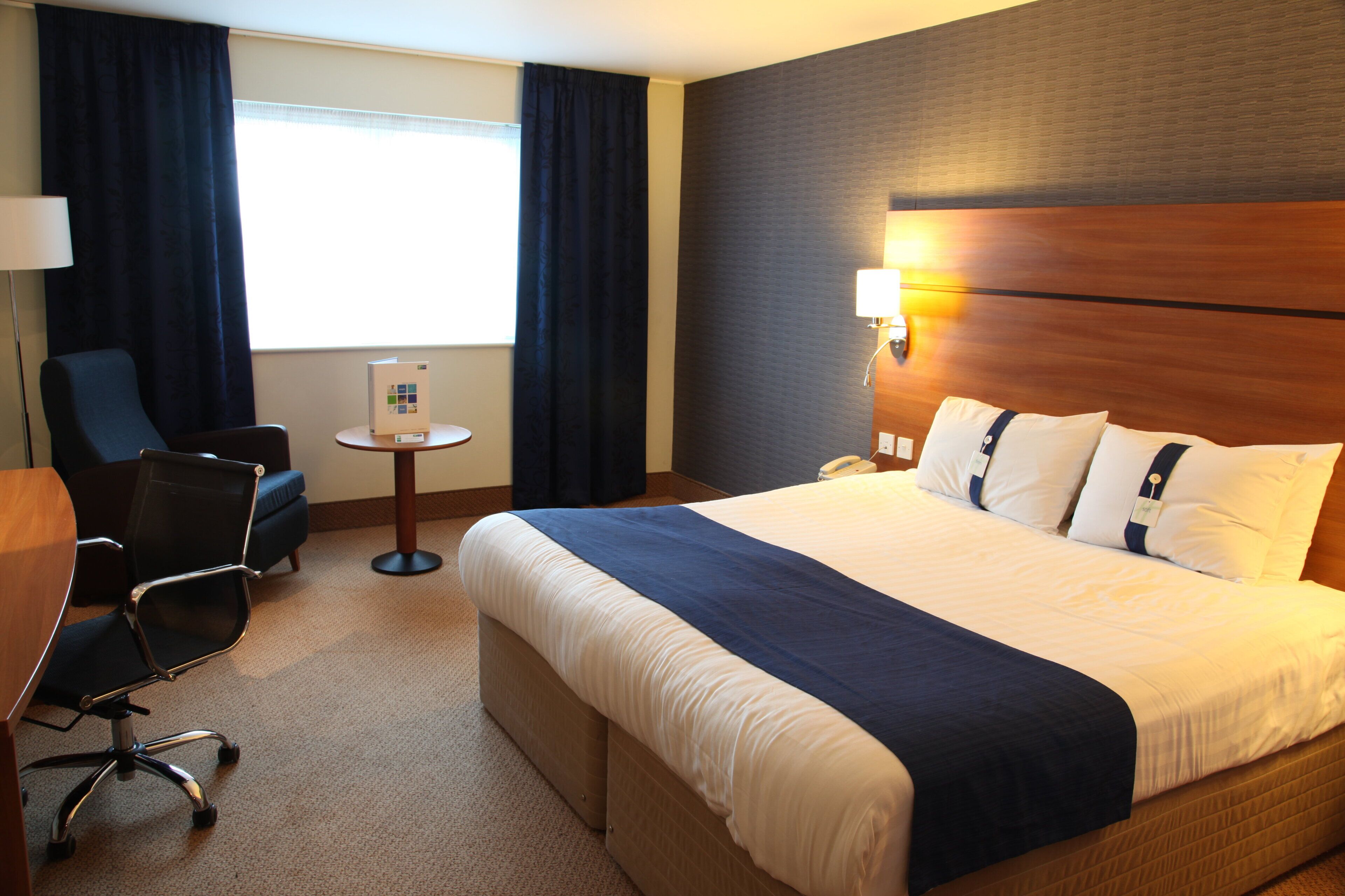 Une chambre d’hôtel avec un grand lit, un bureau, une chaise, une petite table et une lampe.