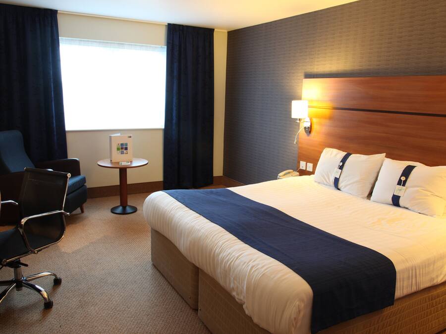 Et hotelværelse med en stor seng, et skrivebord, en stol, et lille bord og en lampe.