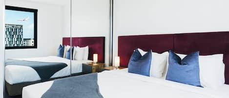 A hotel room with two beds, each with a red headboard, white bedding, and blue pillows. There is a nightstand with a lamp between the beds.