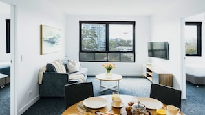 A modern, well-lit living room with a large window, a sofa, a dining table set with plates and cups, and a small side table with a vase of flowers. - Quest Mascot Serviced Apartments (Mascot)